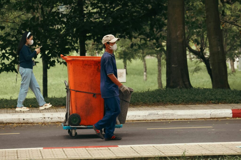 A waste worker pushing a bin cart on a city street while a pedestrian walks by.